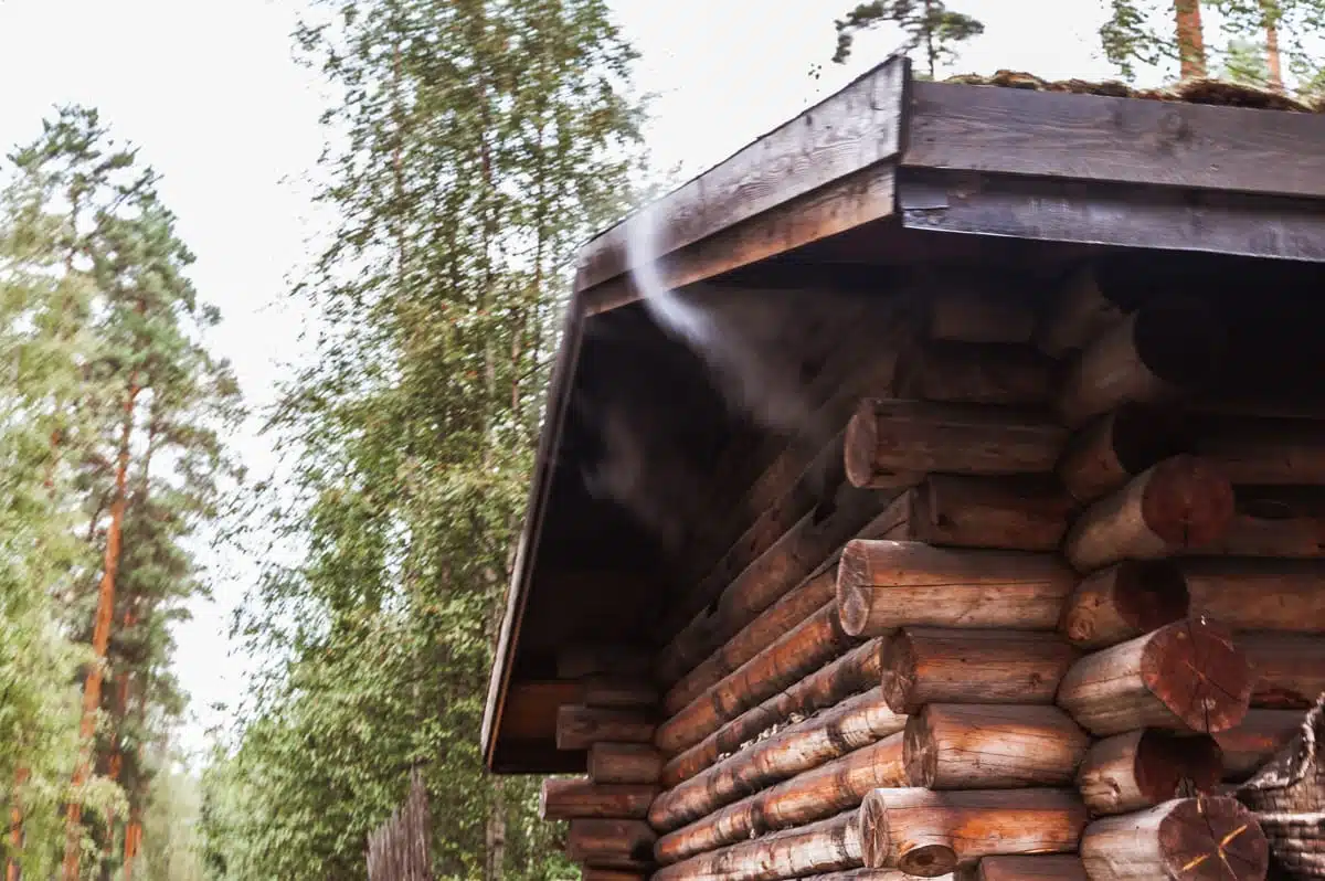 In einem üppigen Wald steht eine rustikale Blockhütte, die an eine traditionelle Rauchsauna erinnert. Unter dem Dachvorsprung steigt sanft Rauch auf, was auf ein warmes Feuer im Inneren hindeutet. Die Bäume ringsum sind hoch und grün, und der Himmel ist leicht bewölkt, was eine friedliche, natürliche Atmosphäre schafft.
