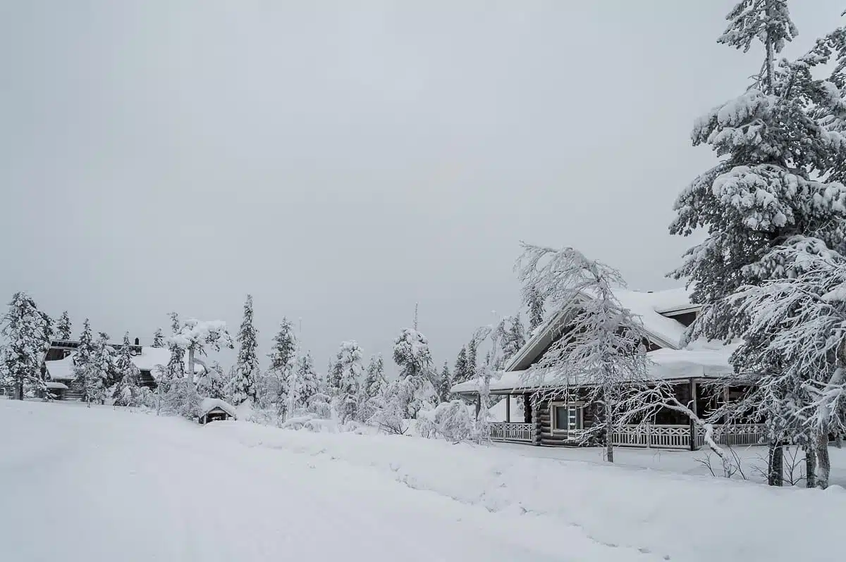 Ein Blockhaus, umgeben von schneebedeckten Bäumen und einer verschneiten Landschaft unter bewölktem Himmel. Schnee bedeckt das Dach, den Boden und alle sichtbaren Flächen und verleiht der Szenerie eine friedliche, winterliche Atmosphäre in Lappland. In der Nähe schlängelt sich neben dem Blockhaus eine schneebedeckte Straße.