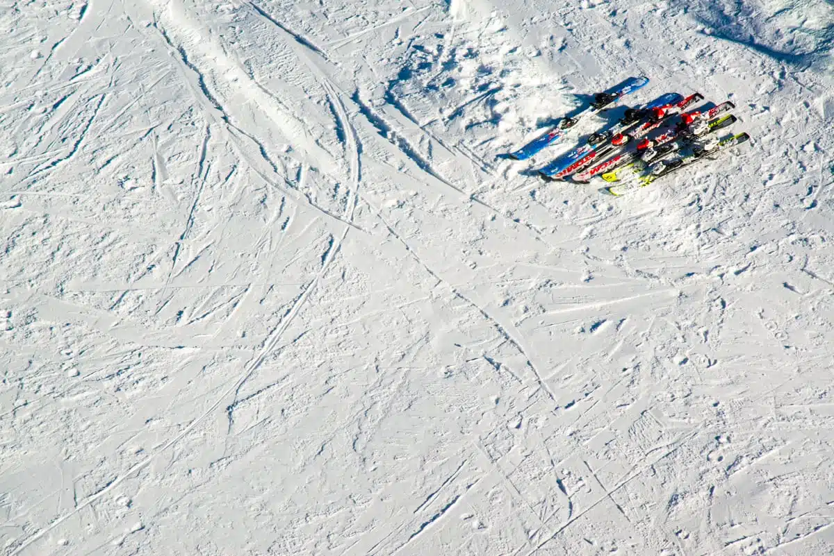 Auf einem weitläufigen, schneebedeckten Hang am Kronplatz, wo das weiße Gelände von zahlreichen Skispuren und Vertiefungen durchzogen ist, liegt eine Ansammlung bunter Skier verlassen herum.