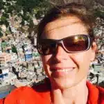 Eine Frau mit braunen Haaren und Sonnenbrille lächelt in die Kamera. Sie trägt eine rote Jacke. Hinter ihr erstreckt sich eine dicht bebaute Stadt am Hang mit bunten Häusern, die dicht aneinander gereiht sind. Das Sonnenlicht spiegelt sich in ihrer Sonnenbrille, und ihr Haar weht im Wind.