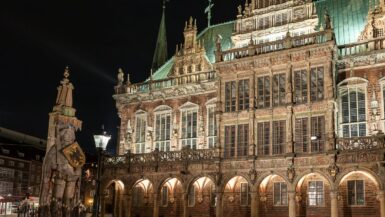 Nachtansicht der kunstvoll verzierten Fassade des Bremer Rathauses mit hohen Bogenfenstern, aufwendigen Schnitzereien und Statuen. Links steht die berühmte Roland-Statue, die ein Schild und ein Schwert hält. Die Szene ist warm beleuchtet, wodurch die architektonischen Details Bremens vor dem dunklen Himmel besonders gut zur Geltung kommen.