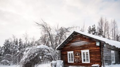 Eine rustikale Holzhütte mit schneebedecktem Dach und Fenstern steht inmitten der verschneiten Landschaft Lapplands. In der Nähe stehen kahle Bäume und ein gekrümmter, schneebedeckter Baum. Im Hintergrund erstreckt sich ein verschneiter Wald unter einem bewölkten, blassen Himmel.