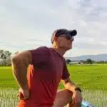 Ein Mann in einem roten Hemd, mit Sonnenbrille und Mütze steht im Freien, lächelt und stützt die Hände in die Hüften. Im Hintergrund sieht man grüne Felder, Berge und einen teilweise bewölkten Himmel. Die Lichtverhältnisse lassen auf den späten Nachmittag oder frühen Abend schließen.