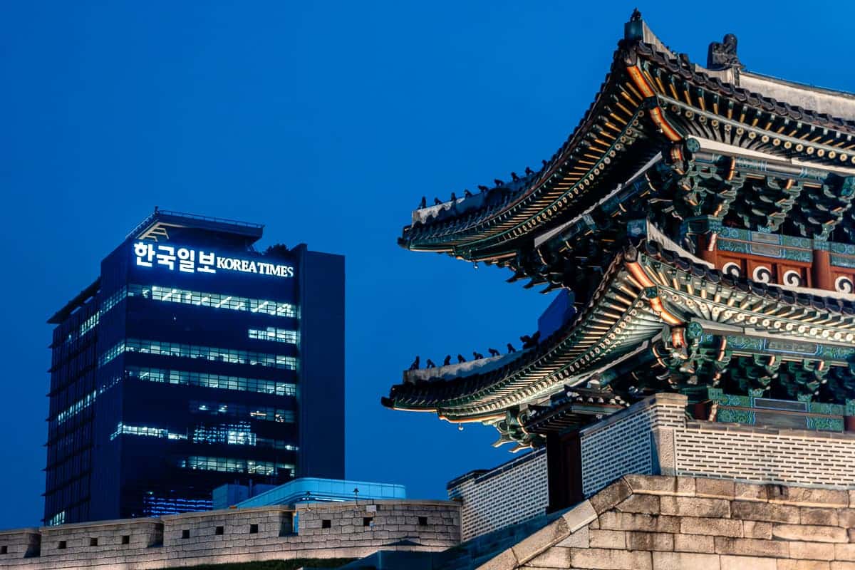 Ausschnitt des Sungnyemun-Tors in Seoul in der Dämmerung, im Hintergrund ein modernes Hochhaus mit Schriftzug "Korea Times".