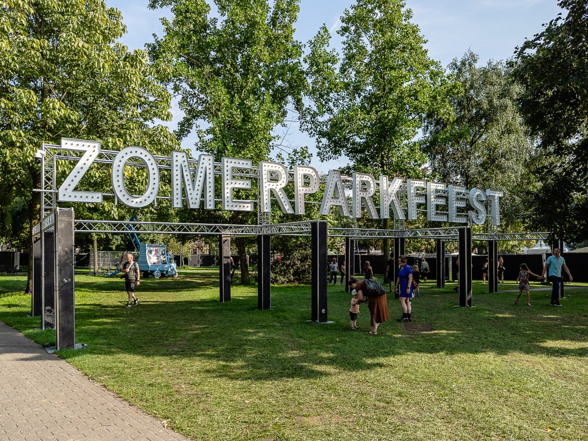 Große Metallbuchstaben bilden das Wort „ZOMERPARKFEEST“ auf einem Rahmen in einem grünen Park mit Bäumen in Venlo. Menschen, darunter Familien und Kinder, spazieren über den Rasen und entspannen sich dort. Es ist sonnig, und im Hintergrund hinter dem Schild ist ein blauer Transporter zu sehen.
