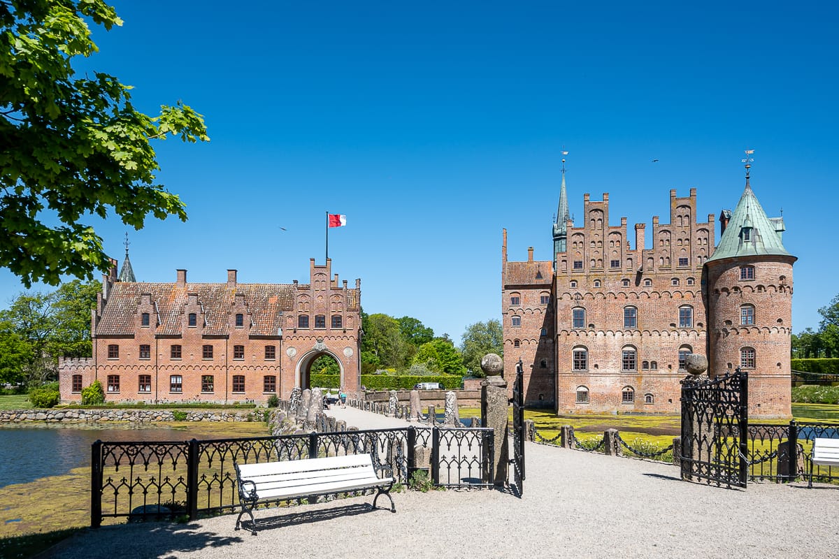 Ein heller, sonniger Tag am Schloss Egeskov in Dänemark mit seinen roten Backsteingebäuden, Türmen, Türmchen und einem Wassergraben. Ein Kiesweg führt über eine Brücke zum Eingang des Schlosses, mit weißen Bänken und einem verzierten schwarzen Geländer im Vordergrund. Über dem Tor weht die dänische Flagge.