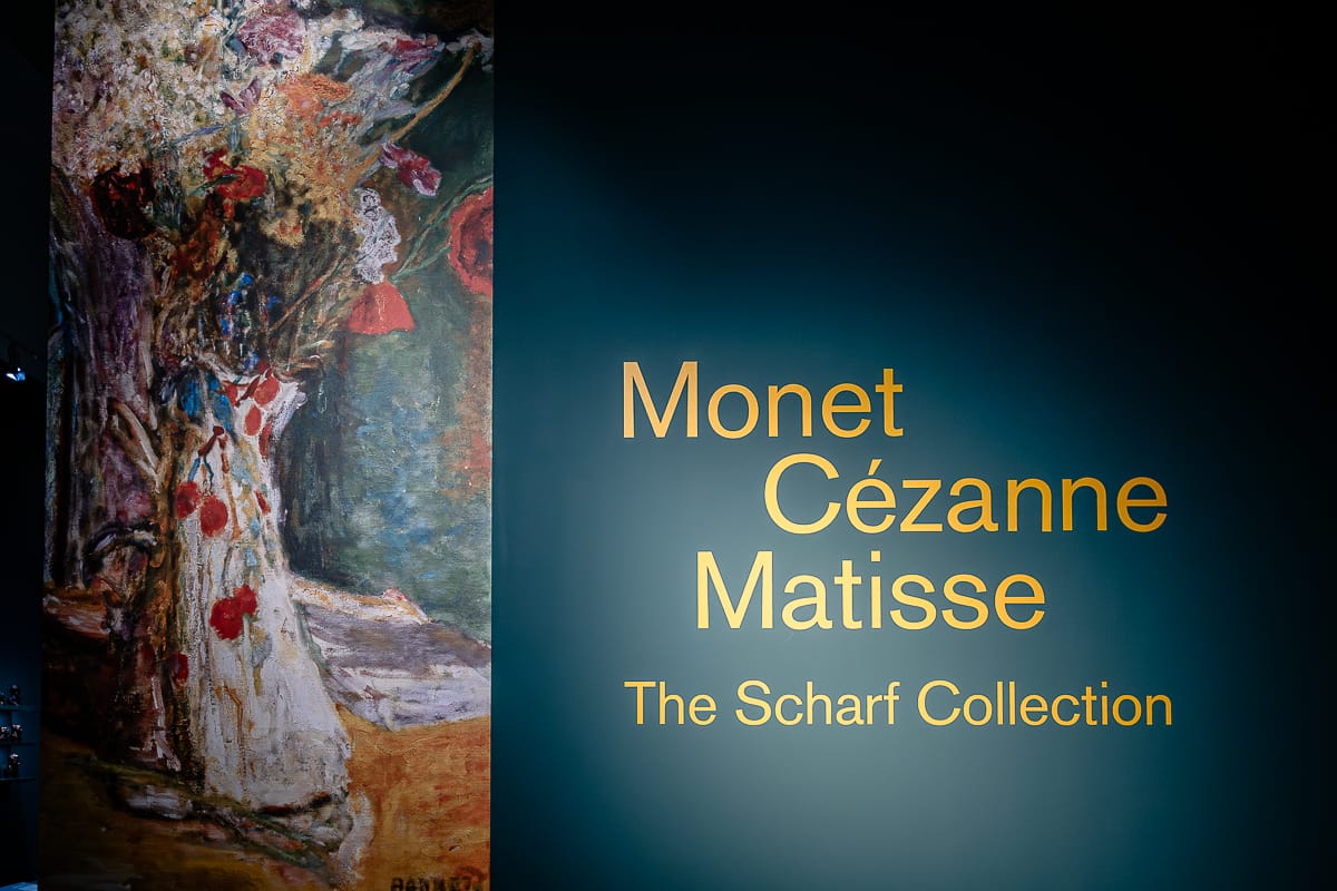 An einer blauen Wand ist neben einem Text, der in fetten goldenen Buchstaben lautet: „Monet, Cézanne, Matisse – Die Scharf-Sammlung“, ein großes Gemälde mit abstrakten Blumenmotiven zu sehen. Die Szene ist sanft beleuchtet, wodurch die Namen und das Kunstwerk besonders hervorgehoben werden.