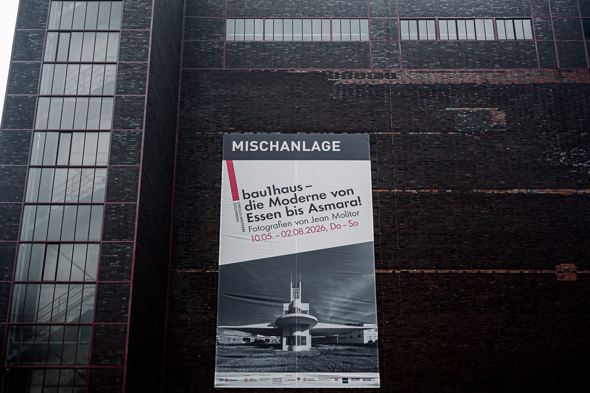Auf einem großen Plakat an einem dunklen Backsteingebäude steht „MISCHANLAGE“ und wirbt für eine Bauhaus-Fotoausstellung von Jean Molitor, auf dem ein Schwarz-Weiß-Foto eines modernistischen Gebäudes zu sehen ist. Die Veranstaltungstermine und der Name des Fotografen sind angegeben. Links sind rote Fensterrahmen zu erkennen.