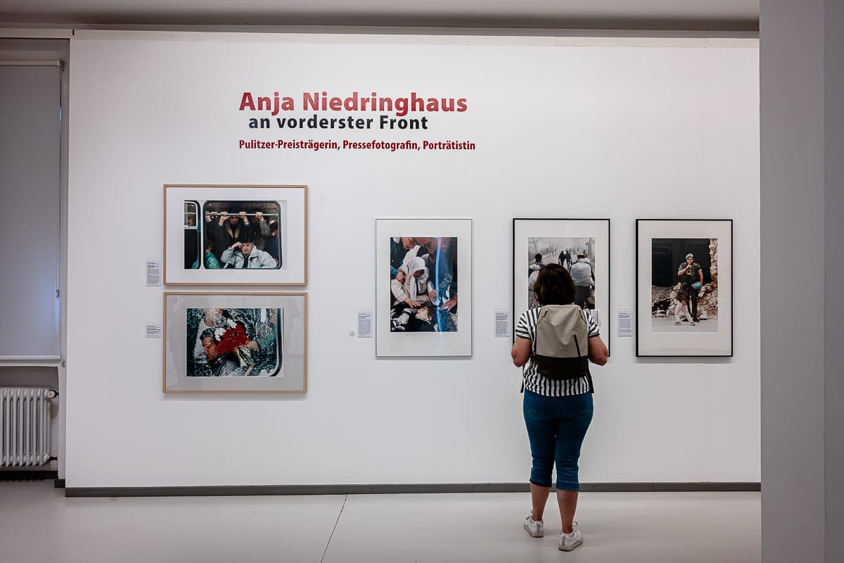 Eine Frau steht vor gerahmten Kriegsfotografien von Anja Niedringhaus in einer Galerie im Rahmen der Anja-Niedringhaus-Ausstellung in Oberhausen. Der Wandtext lautet „Anja Niedringhaus an vorderster Front“ und verweist auf die Ausstellung; neben jedem Foto befinden sich mehrere beschreibende Beschriftungen.