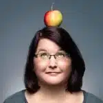 Eine Frau mit dunklem, schulterlangem Haar und Brille blickt zu einem roten und gelben Apfel hinauf, der auf ihrem Kopf balanciert. Sie trägt ein graues Shirt vor einem schlichten grauen Hintergrund. Ihr Gesichtsausdruck ist amüsiert und neugierig.