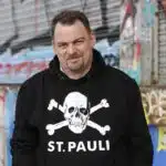 Ein Mann mit kurzen braunen Haaren und einem Spitzbart trägt einen schwarzen Kapuzenpulli mit einem weißen Totenkopf-Motiv und der Aufschrift „ST. PAULI“. Er steht vor einer mit Graffiti übersäten Metallwand und lächelt leicht in die Kamera.