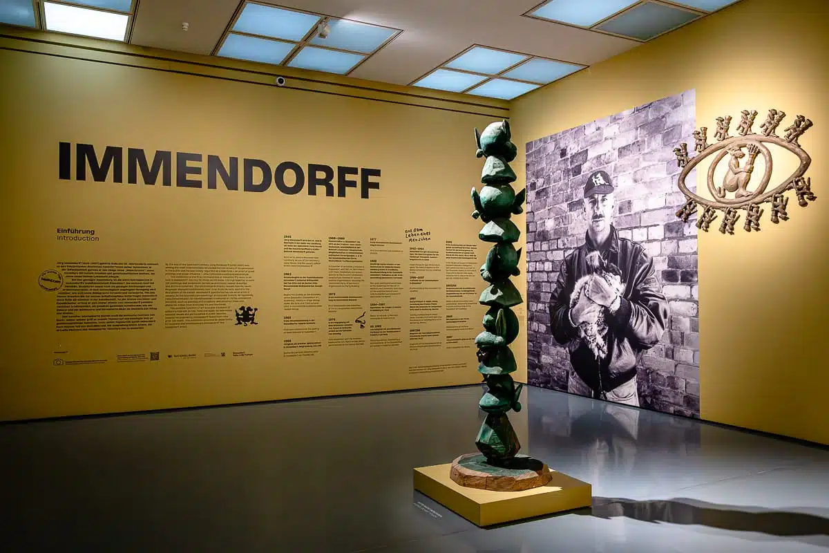 In einer Galerie steht eine hohe, grüne Skulptur aus übereinander gestapelten Figuren vor einer Wand, auf der eine Zeitleiste und eine große Schwarz-Weiß-Fotografie des Künstlers Jörg Immendorff zu sehen sind, der einen Affen im Arm hält; über ihm befindet sich ein augenförmiges Logo.