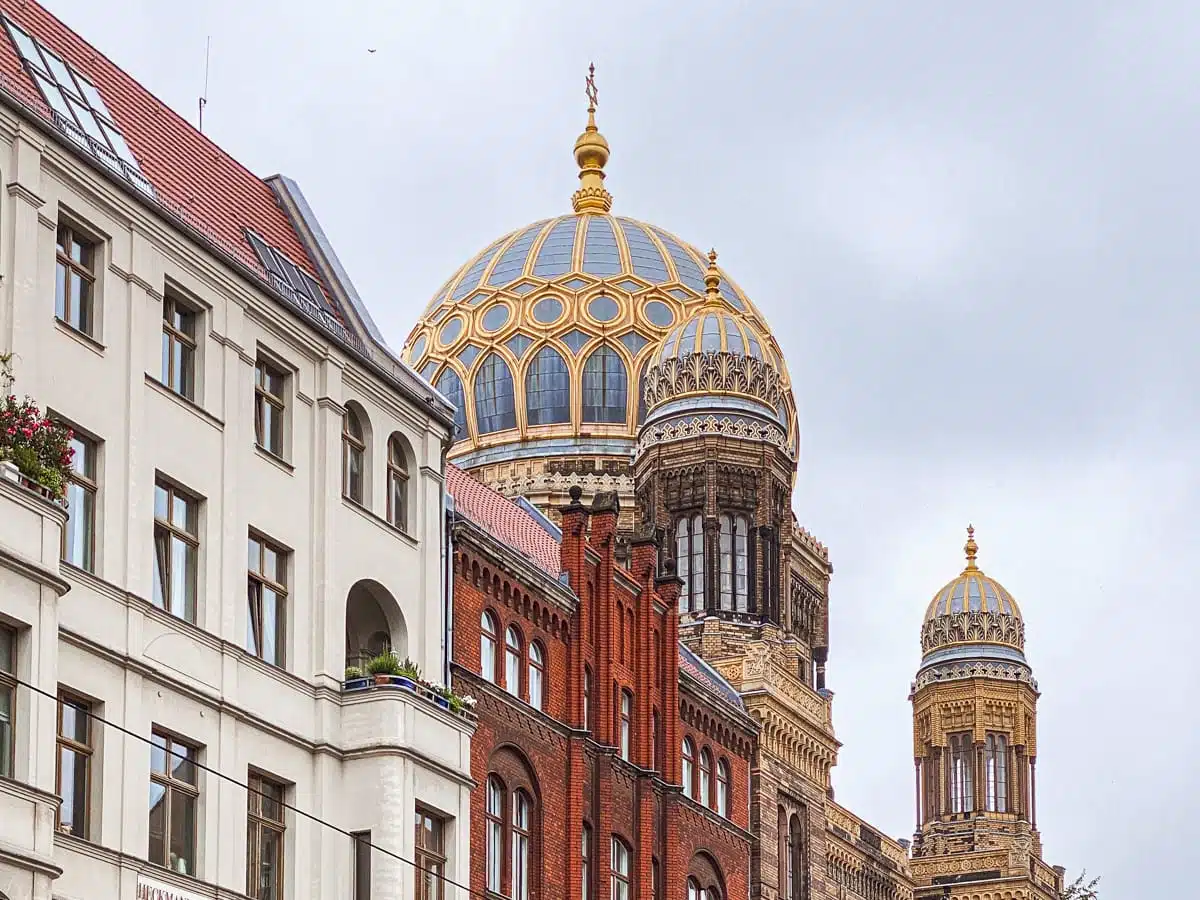 Die Neue Synagoge in Berlin besticht durch eine große, kunstvoll verzierte goldene Kuppel über einer Fassade aus rotem Backstein. Links davon steht unter einem wolkigen grauen Himmel ein weißes Wohnhaus mit rotem Ziegeldach.
