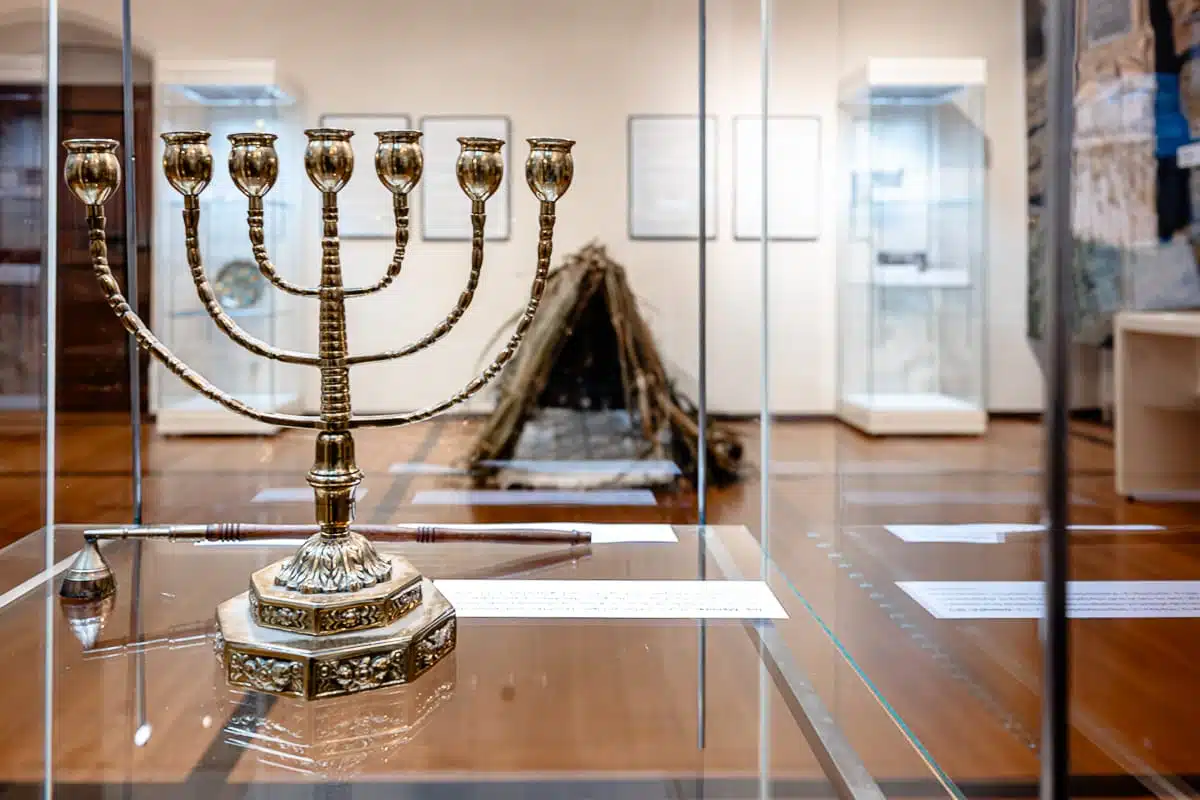 Eine polierte goldene Menora steht auf einer Glasvitrine in einer Museumsausstellung zum Thema „Jüdisches Leben in Delitzsch“. Im verschwommenen Hintergrund erhebt sich zwischen weißen Sockeln und gerahmten Dokumenten an den Wänden der Galerie eine hölzerne, Tipi-ähnliche Konstruktion.