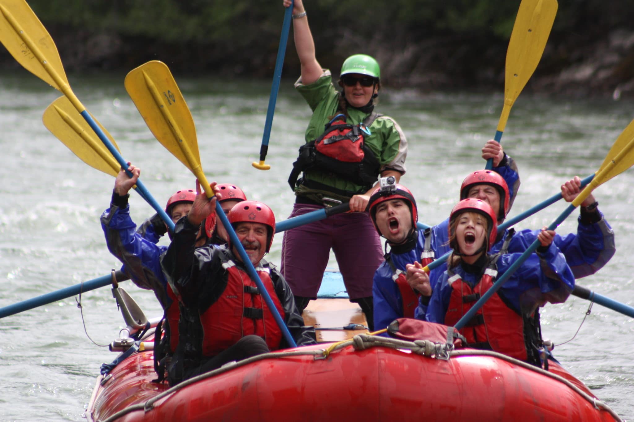 Eine Gruppe von sieben Personen in rot-blauer Ausrüstung feiert ihr Rafting-Abenteuer auf einem roten Schlauchboot im Fluss. Triumphierend halten sie gelbe Paddel in die Höhe, in der Mitte steht eine Person mit grünem Helm und streckt einen Arm in die Höhe. Im Hintergrund ist verschwommenes Grün am Flussufer zu sehen.
