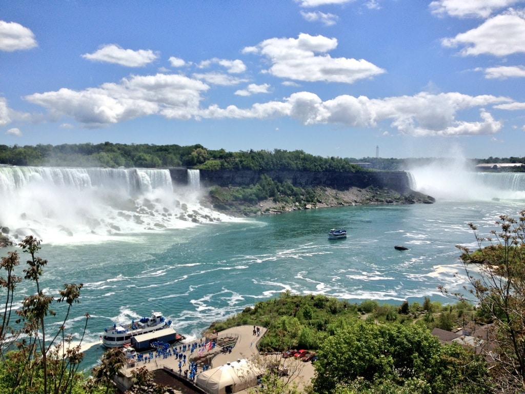 Eine malerische Aussicht auf die Niagarafälle unter einem blauen Himmel mit vereinzelten Wolken. Zwei Wasserfälle stürzen in den darunterliegenden Fluss, wo zwei Ausflugsboote in der Nähe der Wasserfälle fahren. Üppiges Grün umgibt die Landschaft, im Vordergrund sind Bäume und Büsche zu sehen. Touristen versammeln sich am Flussufer.