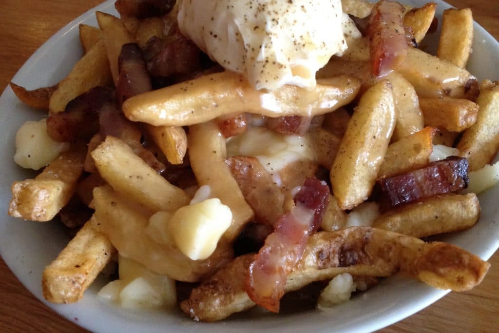Eine Schüssel Breakfast Poutine in Saskatoon, Saskatchewan. Sie besteht aus goldbraunen Pommes Frites, die mit einer großzügigen Portion Käsebruch belegt und mit einer reichhaltigen, würzigen Soße übergossen werden. Knusprige Speckstücke sind obenauf verteilt und sorgen für zusätzlichen Geschmack und Knusprigkeit. Ein porchiertes Ei mit schwarzem Pfeffer krönt das Ganze.