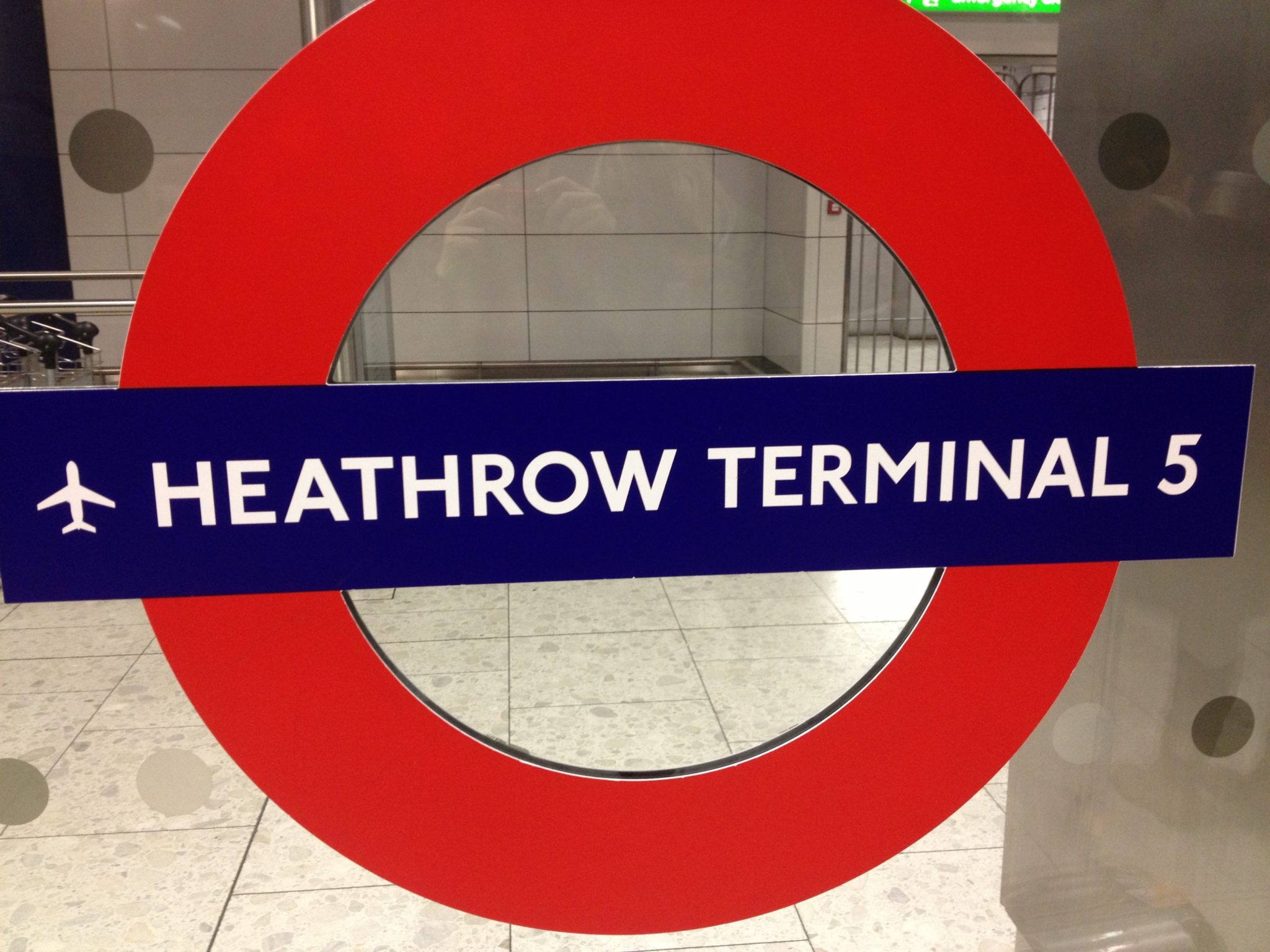 Ein Schild zeigt das ikonische Rundschild der Londoner U-Bahn. Auf dem blauen Balken steht in strahlend weißen Buchstaben „Heathrow Terminal 5“, begleitet von einem Flugzeugsymbol. Vor dem Hintergrund eines gefliesten Bodens und eines Teils der Innenansicht der Station deutet es auf Reiseabenteuer auch im Winter in London hin.