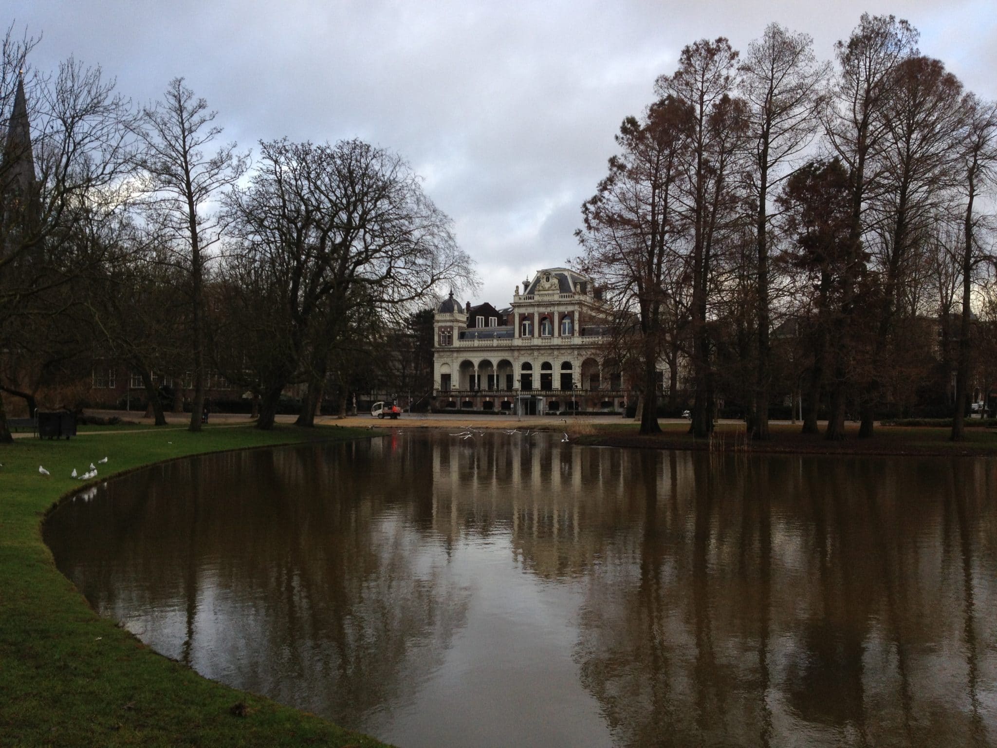 Vondelpark - Blick auf ein historisches Gebäude im Hintergrund mit hochen Bäumen links und rechts, großem Teich davor, an dem links Enten am Rand stehen.