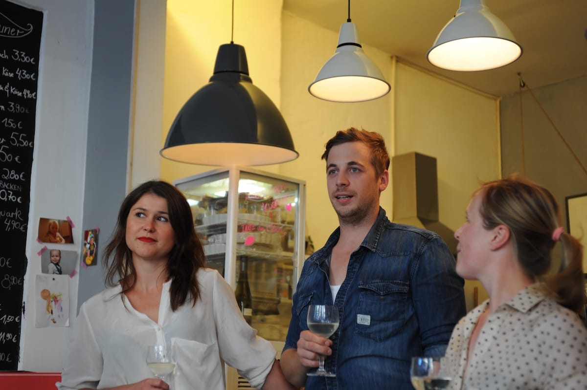 Drei Personen stehen in einem Haus und halten Weingläser. Die Frau links in einer weißen Bluse hält den Arm des Mannes in einem Jeanshemd. Auf der rechten Seite steht eine blonde Frau. Hängelampen und eine Tafel geben die Stimmung vor.