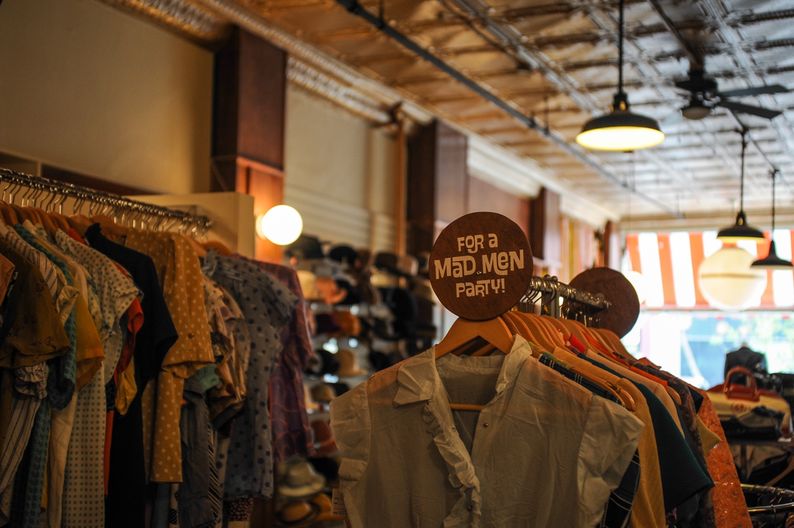 Gezeigt wird ein Vintage-Bekleidungsgeschäft mit Ständern voller bunter Hemden und einem runden Schild mit der Aufschrift „Für eine Mad Men-Party!“. Das Geschäft mit dem Namen „Rhymes With Orange“ hat eine Retro-Ästhetik mit warmer Beleuchtung, einer gemusterten Blechdecke und großen Fenstern, die mit gestreiften Markisen geschmückt sind.