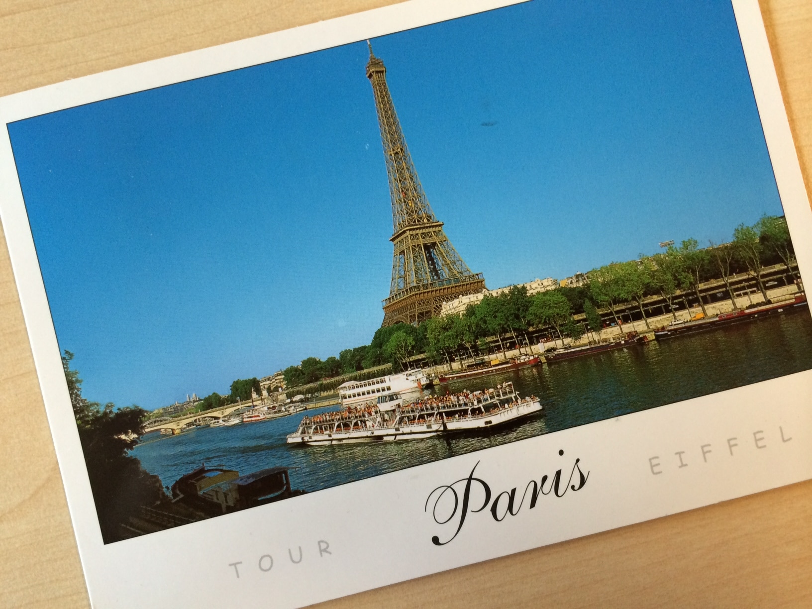 Postkarte von Paris mit dem berühmten Eiffelturm unter einem klaren blauen Himmel. Im Vordergrund fließt die Seine anmutig dahin, ein Ausflugsboot voller Menschen fährt vorbei. Bäume säumen das Flussufer, und unten ist „Tour Eiffel“ aufgedruckt, was die helle, sonnige Atmosphäre von Paris einfängt.