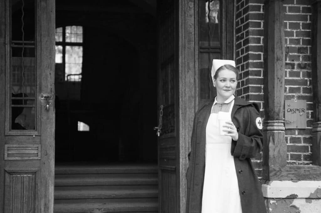 Eine Krankenschwester in weißer Uniform und Haube steht mit einer Tasse in der Hand vor einem Backsteingebäude. Sie trägt einen dunklen Mantel und eine Armbinde des Roten Kreuzes. Das Schwarz-Weiß-Foto entstand während eines Fotoworkshops und zeigt offene Türen, hinter denen Treppen und Bogenfenster zu sehen sind.