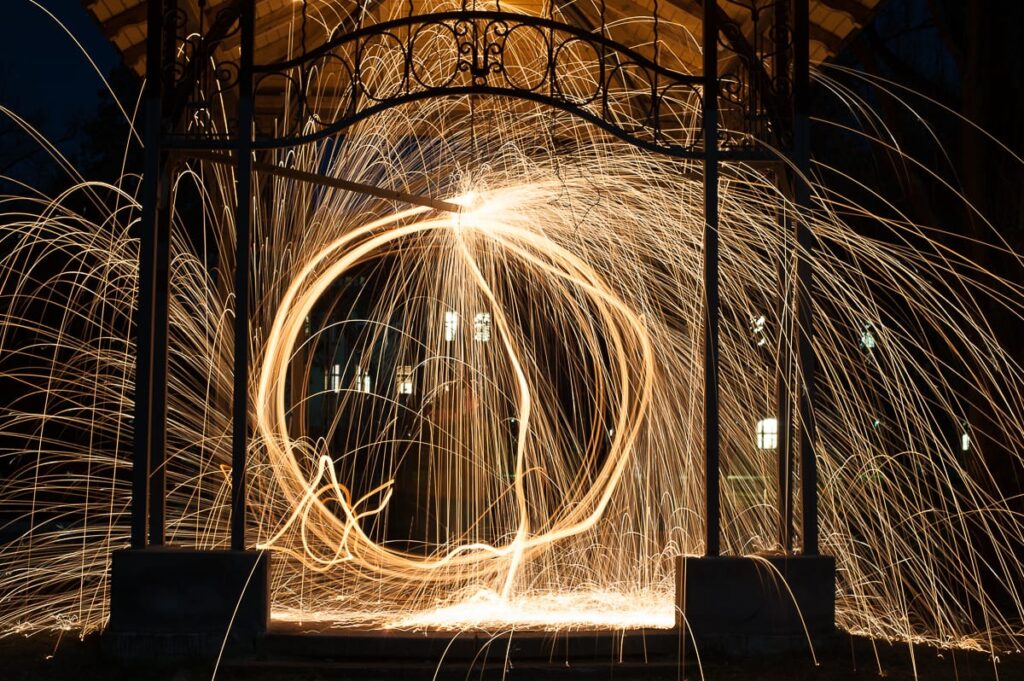 Ein Langzeitbelichtungsfoto aus einem Fotoworkshop zeigt das Spinnen von Stahlwolle unter einem Pavillon bei Nacht, wobei helle, wirbelnde gold-orangefarbene Funkenspuren das dunkle Metallgerüst beleuchten und im Hintergrund leuchtende Fenster zu sehen sind.