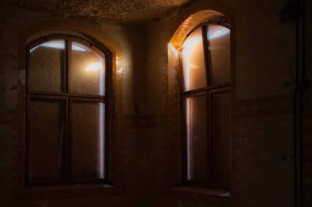 Zwei große Bogenfenster in einem schwach beleuchteten, verlassenen Raum. Schwaches Sonnenlicht fällt durch das schmutzige Glas und wirft sanftes orange-weißes Licht auf die alten Backsteinwände – die ideale Kulisse für einen stimmungsvollen Fotoworkshop, der Licht und Atmosphäre erforscht.