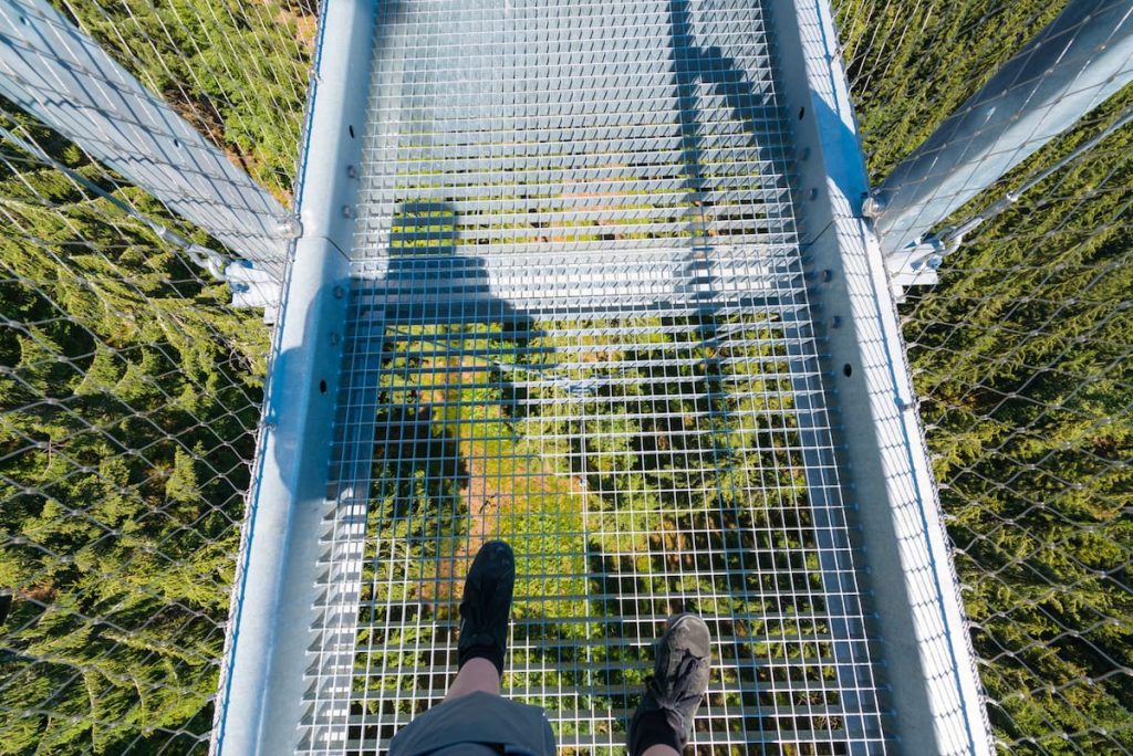 Eine Person steht auf einer Metallgitter-Hängebrücke hoch über einem Wald. Der Blick fällt auf ihre Füße und ihren Schatten, weit unten sind grüne Baumkronen durch das offene Gitter der Brücke zu sehen.
Foto: Simon Bierwald/Indeed Photography