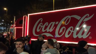 Ein hell erleuchteter Coca-Cola-Weihnachtstruck steht nachts draußen am CentrO Oberhausen und ist von einer fröhlichen Menschenmenge umgeben. Der Truck, geschmückt mit weißen und gelben Lichtern, die das rote Coca-Cola-Logo hervorheben, zieht Menschen jeden Alters in Winterkleidung an, einige von ihnen werden von aufgeregten Kindern begleitet.