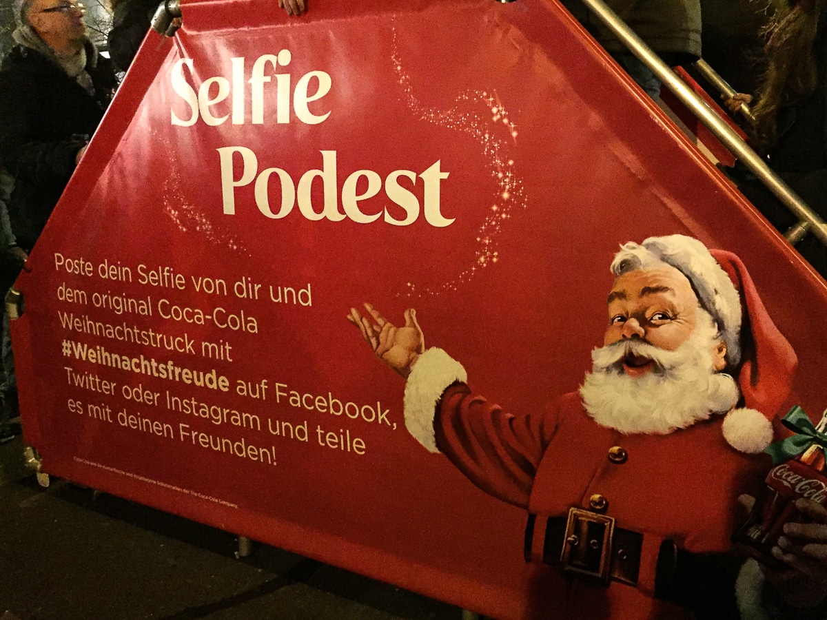 Ein festliches Schild in deutscher Sprache zeigt den Weihnachtsmann vor einem Weihnachtstruck-Hintergrund und wirbt für ein Selfie-Podest. In seinem ikonischen roten Anzug gestikuliert er spielerisch, um zum Teilen von Selfies in sozialen Medien mit dem Hashtag #Weihnachtsfreude aufzufordern und hebt dabei Plattformen wie Facebook, Twitter und Instagram hervor.