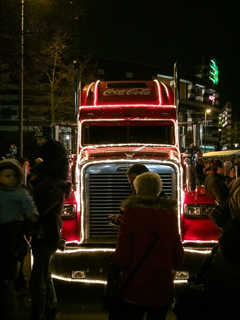 Ein hell erleuchteter Weihnachtstruck, ein Coca-Cola-Truck, geschmückt mit Lichterketten, steht nachts geparkt. Menschen versammeln sich am CentrO Oberhausen um ihn herum, einige machen Fotos. Im Hintergrund sind hohe Gebäude mit beleuchteten Schildern zu sehen. Die Atmosphäre ist festlich, die Menschen sind in Winterkleidung gekleidet.