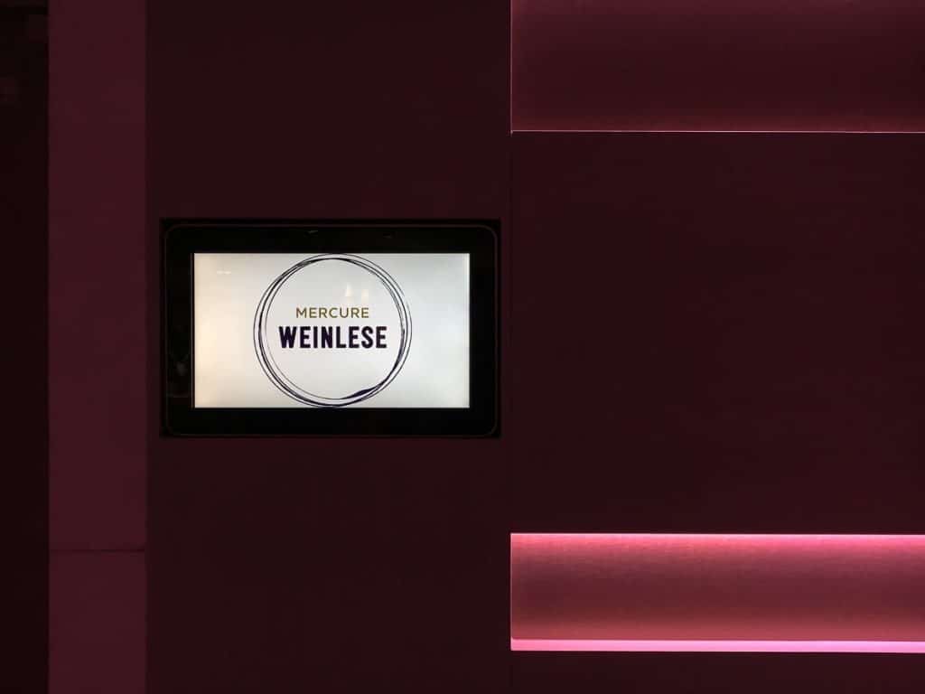 Ein Bildschirm zeigt "Mercure Weinlese" in einem kreisförmigen Design auf einer dunklen Wand mit horizontalen, rosa beleuchteten Akzenten. Die kastanienbraune und rosafarbene Farbgebung schafft eine moderne, stilvolle Atmosphäre, die die Eleganz der Mercure Weinlese unterstreicht.