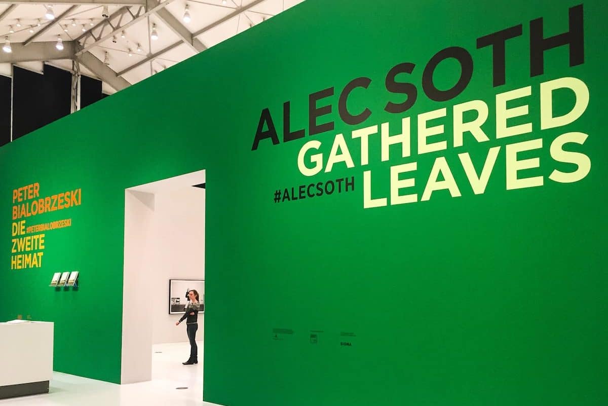 Ausstellungseingang mit leuchtend grünen Wänden und großen Texten: „ALEC SOTH GATHERED LEAVES“ und „PETER BIALOBRZESKI DIE ZWEITE HEIMAT“ in Gelb und Weiß. Eine Person im Inneren scheint die ausgestellte Kunst zu betrachten. Die Decke hat ein geometrisches Gittermuster.