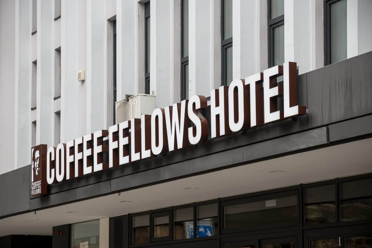 Das Bild zeigt das elegante Äußere des Coffee Fellows Hotels mit einem fettgedruckten, weißen Schriftzug vor einem dunklen Hintergrund. Große Fenster unterstreichen das moderne Design, während sich ein Kaffeetassensymbol nahtlos in das Schild einfügt und die unverwechselbare Marke „Coffee Fellows“ verkörpert.