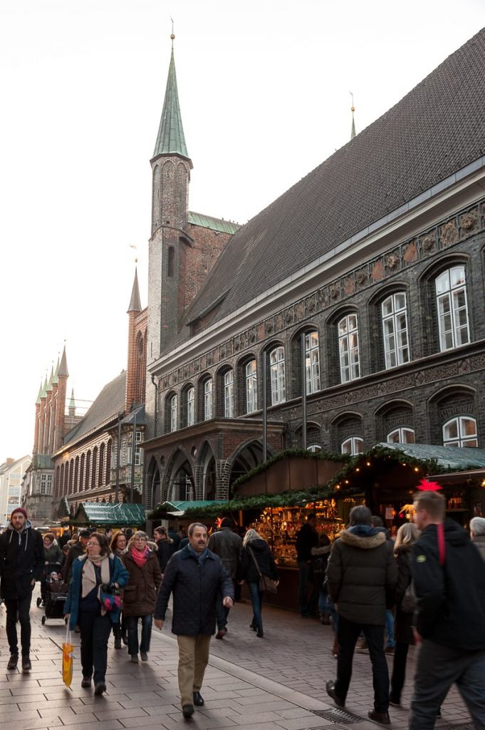 Menschen schlendern über einen lebhaften Weihnachtsmarkt in der charmanten Breiten Straße von Lübeck. Festlich geschmückte Stände säumen den Kopfsteinpflasterweg. Ein historisches Gebäude mit komplexer Architektur und einem hohen Turm sticht hervor. Käufer, eingemummelt in Winterkleidung, tragen zur geschäftigen Atmosphäre bei.