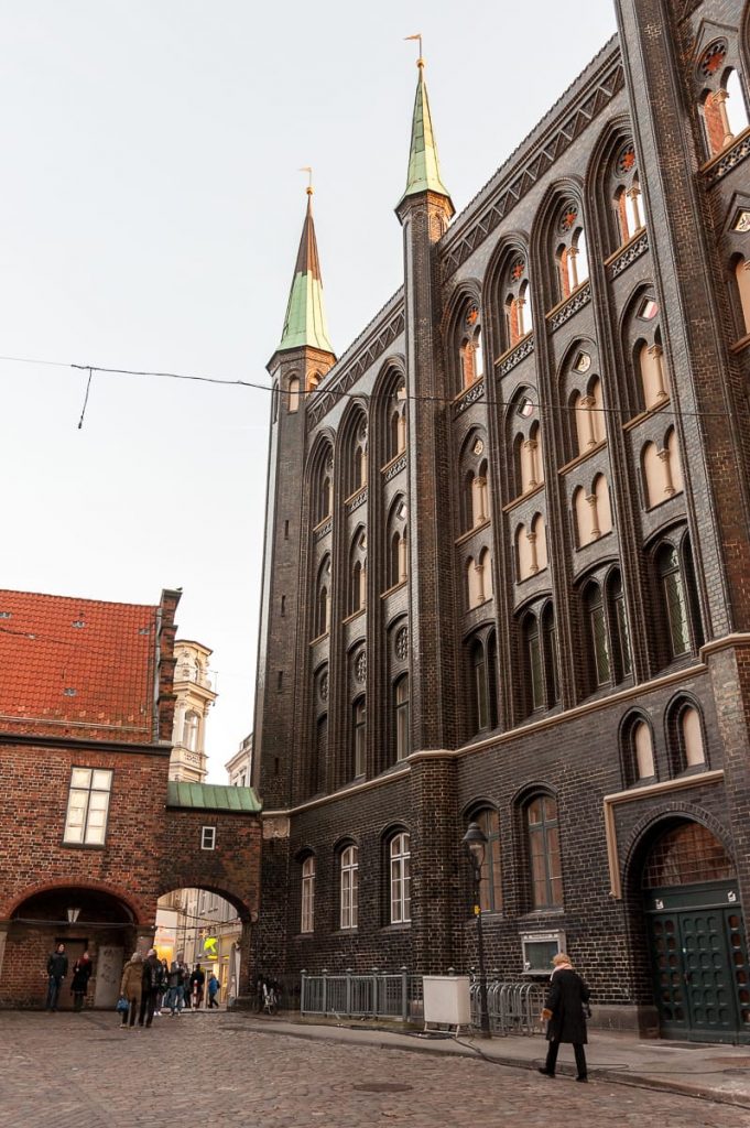 Eine Kopfsteinpflasterstraße in Lübeck (Breite Straße) führt zu einem großen Backsteingebäude im gotischen Stil mit zwei hohen, spitzen Türmen, die von grünen Spitzen gekrönt werden. Mehrere Bogenfenster säumen die Fassade. Eine kleine Gruppe von Menschen schlendert in der Nähe umher, während ein paar Fußgänger über die Szenerie verstreut sind.