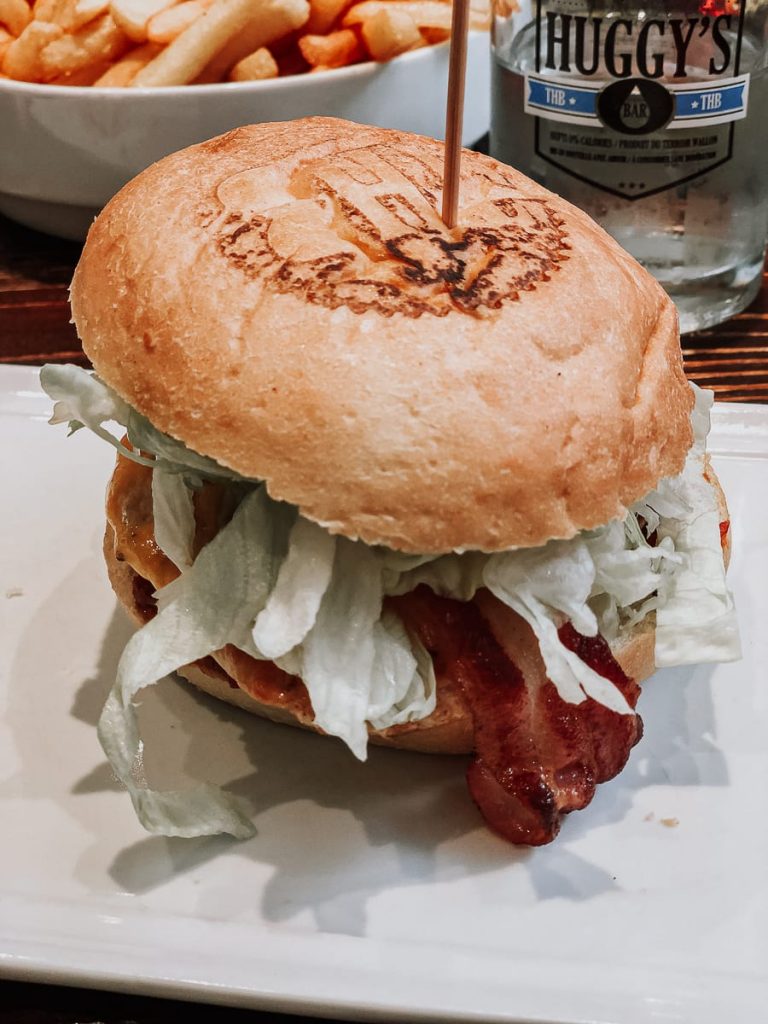 Ein Burger mit getoastetem Brötchen, Salat, Käse und knusprigem Speck auf einem weißen Teller, zusammengehalten von einem Spieß. Im Hintergrund liegen Pommes in einer Schale neben einem Glas, das mit „Huggy’s“ von Lüttich verziert ist.