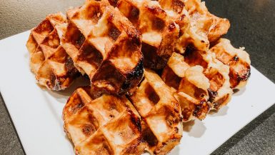 Ein Stapel goldbrauner, karamellisierter Lütticher Waffeln liegt auf einem weißen Teller. Die Waffeln haben eine knusprige Außenseite mit sichtbaren Zuckerflecken und unebenen Rändern, was ihr hausgemachtes, rustikales Aussehen auf einer dunklen Arbeitsplatte unterstreicht.