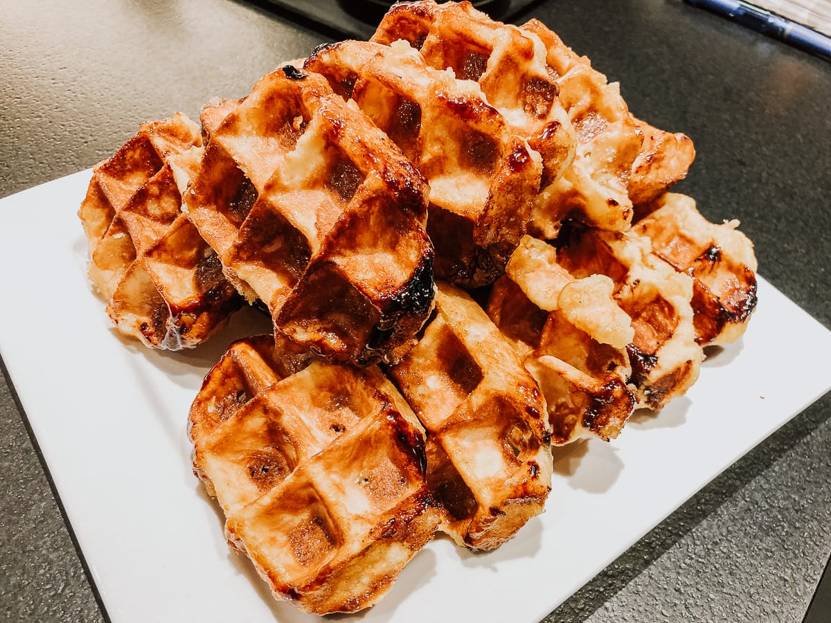Ein Stapel goldbrauner, karamellisierter Lütticher Waffeln liegt auf einem weißen Teller. Die Waffeln haben eine knusprige Außenseite mit sichtbaren Zuckerflecken und unebenen Rändern, was ihr hausgemachtes, rustikales Aussehen auf einer dunklen Arbeitsplatte unterstreicht.