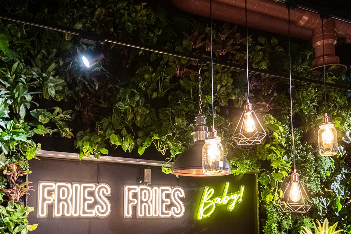 Fries Fries baby-Leuchtschrift im Erdgeschoss, umgeben von vielen Pflanzen, davor einige Lampen im Industriestil.