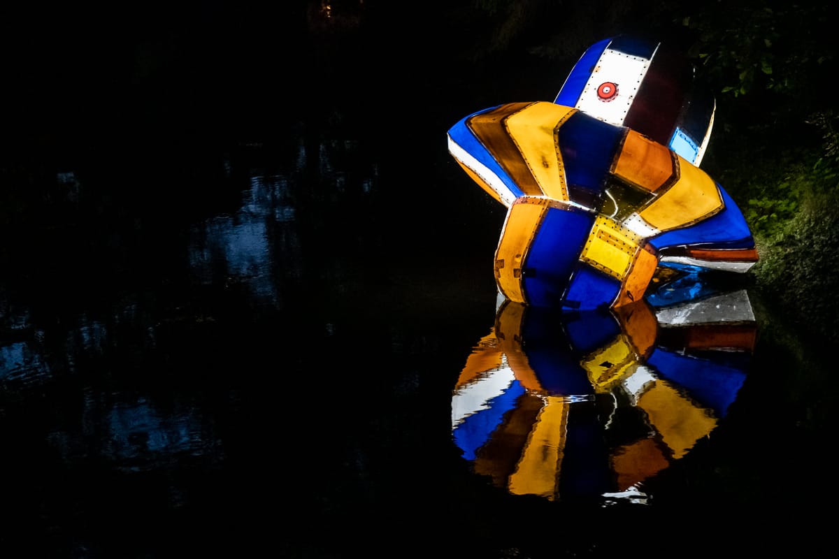 Eine farbenfrohe, abstrakte Skulptur mit geometrischen Formen in Blau, Gelb, Weiß und Orange wird vor einem dunklen Hintergrund beleuchtet. Als Teil der Ausstellung Lichtparcours 2020 steht sie neben einem reflektierenden Gewässer und spiegelt dessen lebendige Farben und Muster auf der ruhigen Oberfläche wider.