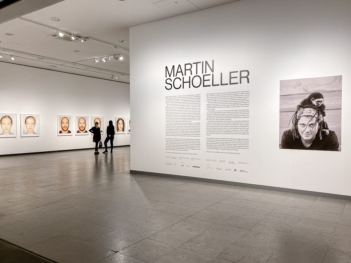 Eingang zur Werkschau von Martin Schoeller im NRW-Forum. Blick auf sein Foto und Biografie, dahinter die Serie "Identicals" mit zwei Besuchern.
