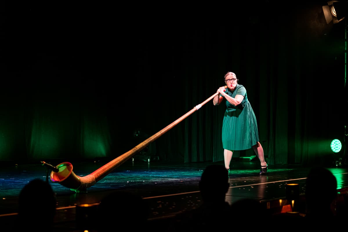 Rosemie bläst gleich in ihr Alphorn, Lippen sind gespitzt. Sie hat ein grünes Kleid an.