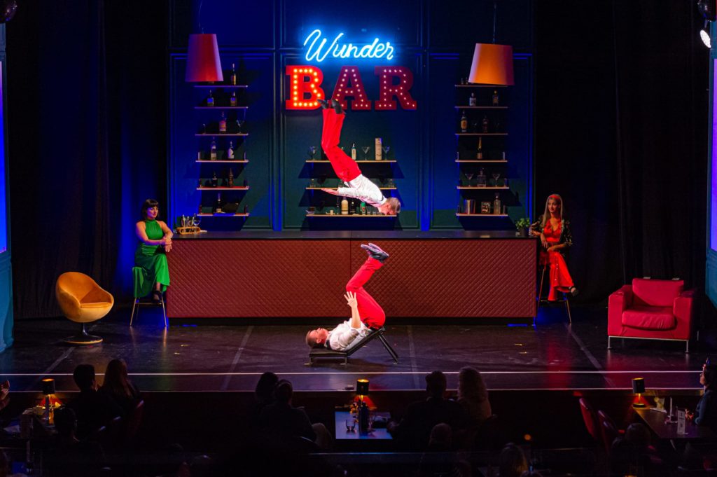 Das Duo Fabulous in der Mitte der Bühne, links und rechts der Bar sitzen die Dame in Grün und die Barbesitzerin der WunderBar. Das Duo Fabulous wirbelt einander durch dei Luft (Ikarische Spiele, einer liegt auf einer Art Liegestuhl, der andere befindet sich gerade in der Luft)