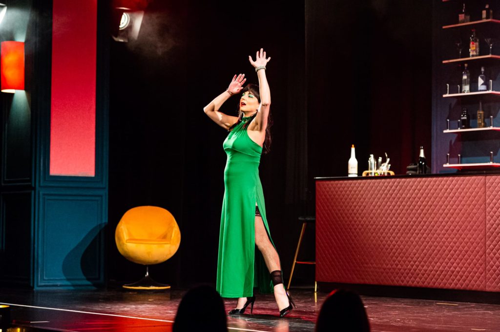 Ava, die Dame in Grün, steht mit einem Ausfallschritt in der WunderBar. Durch den hohen Schlitz im Kleid wird das linke Bein sichtbar, in schwarzen Nylons. 