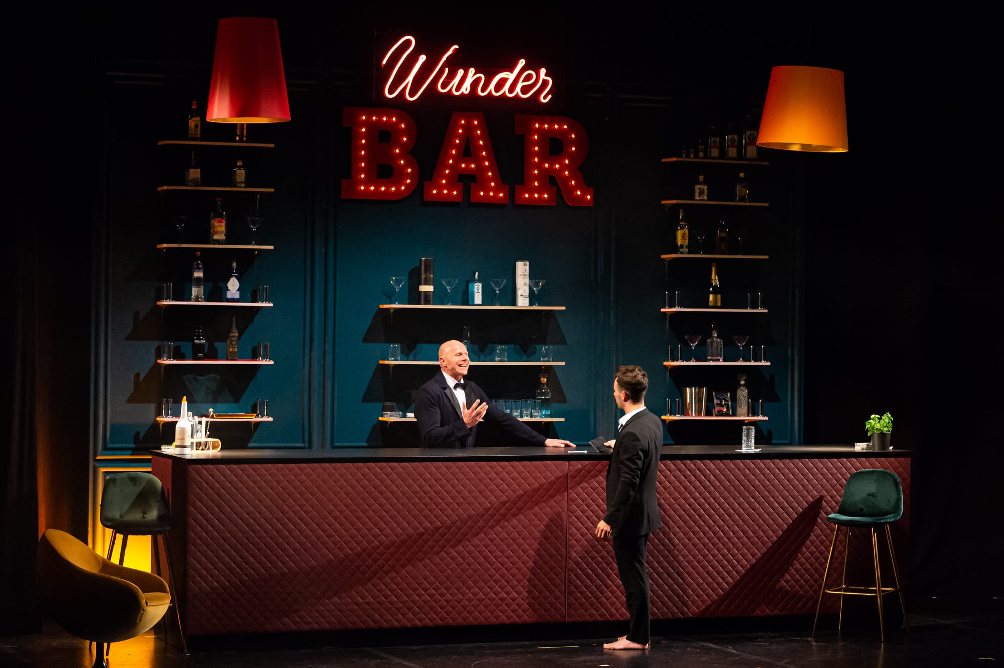 Ein Barkeeper unterhält sich mit einem Kunden an der Theke einer stilvollen Bar. Die Bar verfügt über beleuchtete Regale mit Flaschen und Gläsern. Darüber steht auf einem roten Neonschild „Wunder Bar“. Die Szene ist mit hängenden gelben Lichtern warm beleuchtet und an den Seiten sind Plüschsitze zu sehen.