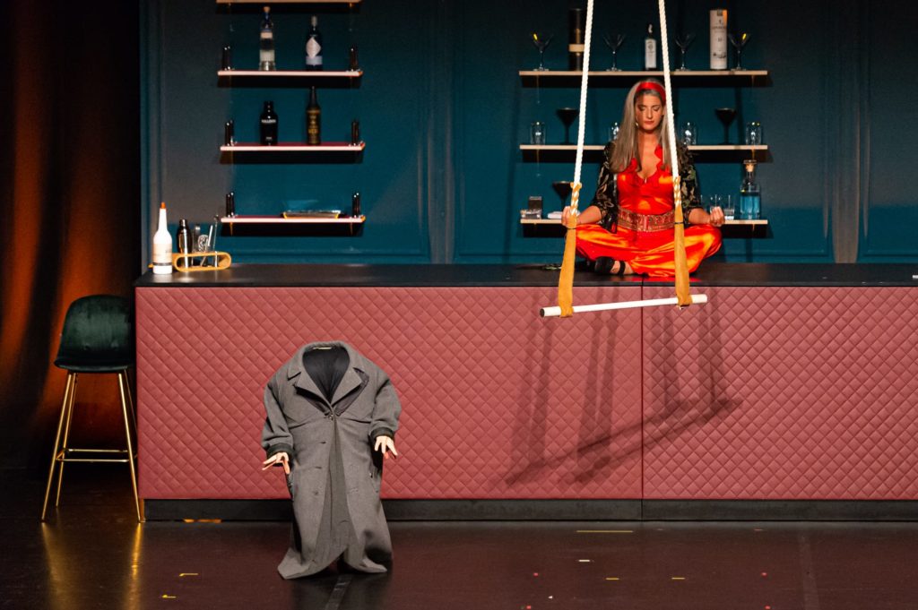 Die Sängerin sitzt im Schneidersitz auf dem Tresen der WunderBar, Hände nach oben, Yogapose, meditierend (Augen geschlossen), vor ihr hängt eine Trapezstange, ein "kleiner Mann" ohne Kopf, nur Hände, grauer Mantel steht links. 