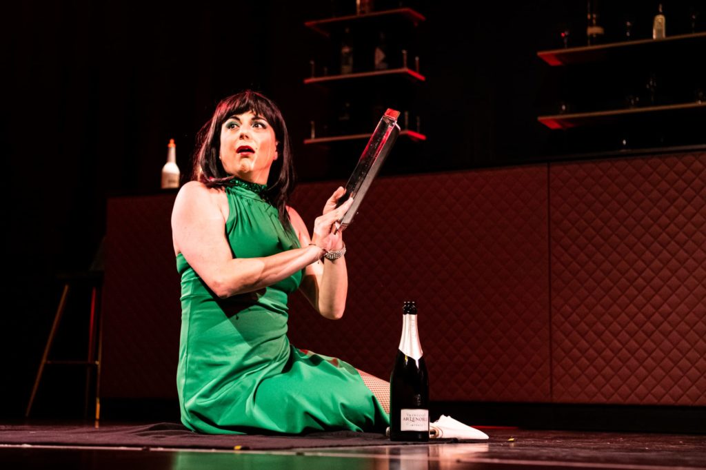 Ava, die Dame in Grün, sitzt im Kleid auf dem Fußboden der WunderBar, neben ihr steht eine Flasche Champagner, sie hat etwas längliches, metallisch-glänzendes in der Hand und schaut etwas überrascht ins Publikum. 